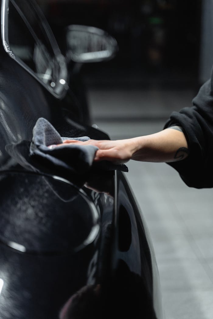 Close-up of a person's hand cleaning a shiny black car with a microfiber cloth in a garage.